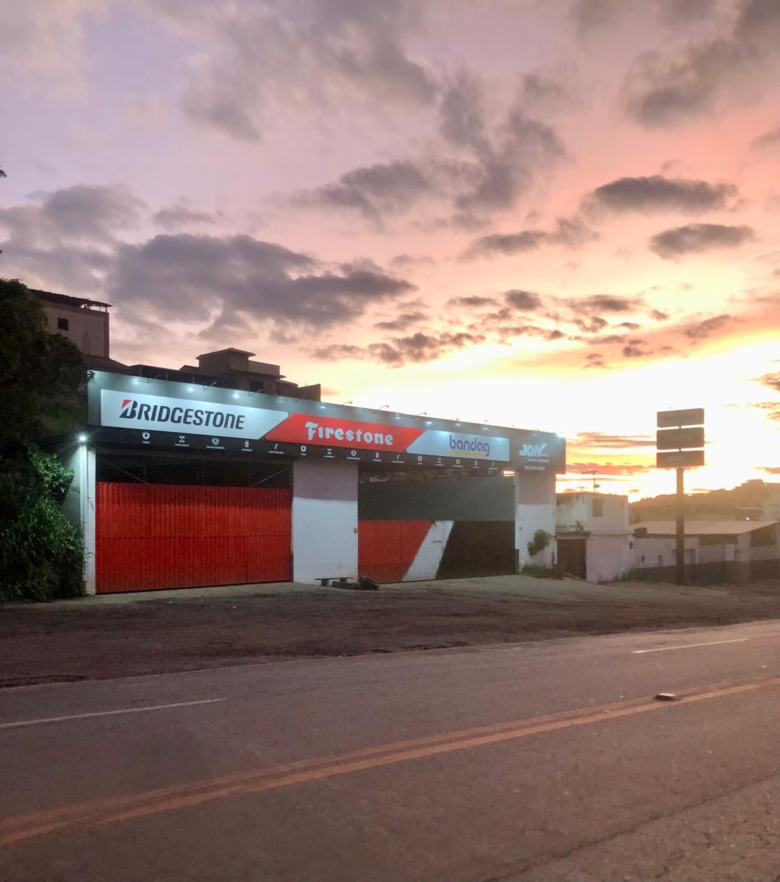 Fachada da JOAZ Pneus iluminada ao pôr do sol, à beira da BR-040 em Conselheiro Lafaiete